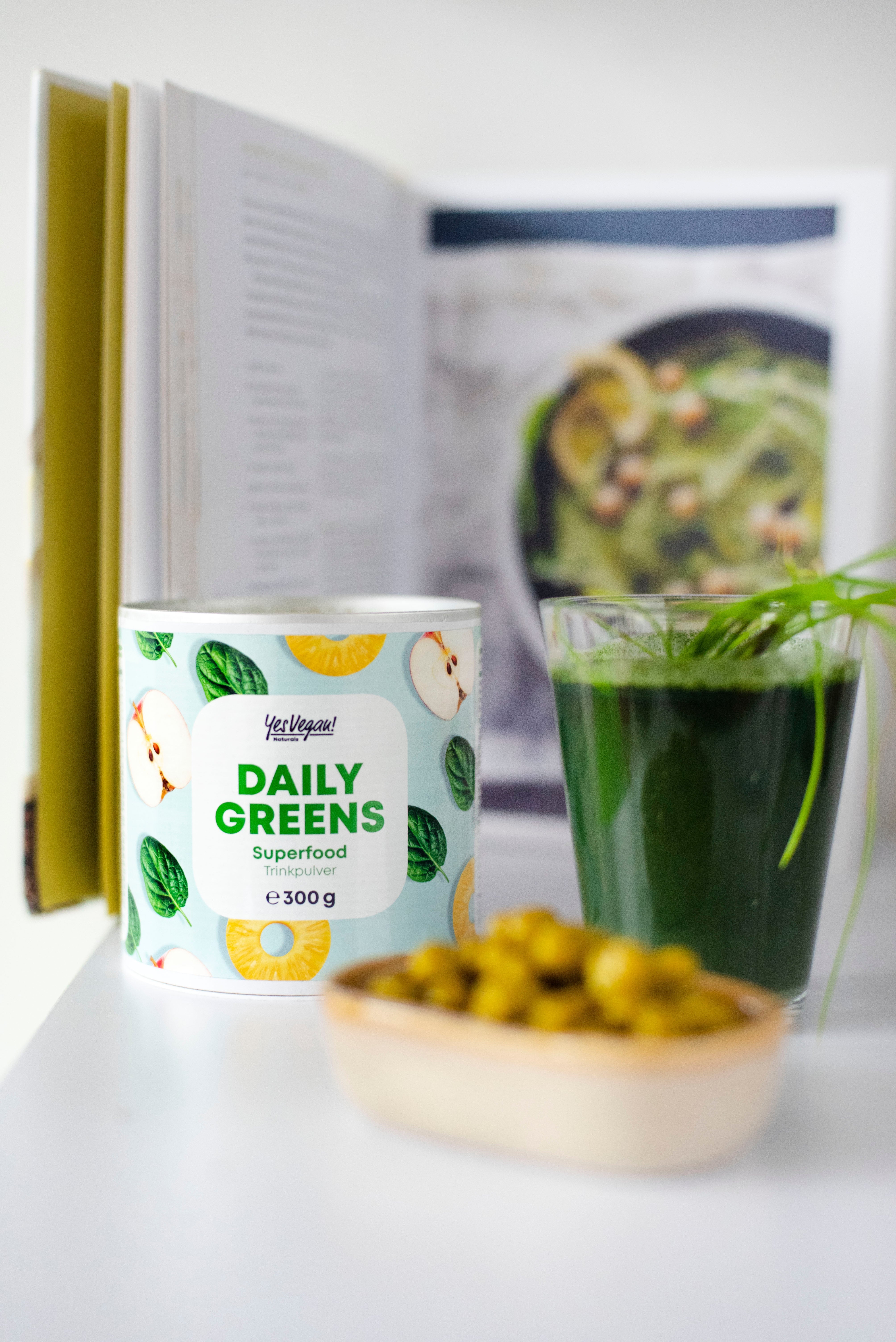 Yes Vegan Daily Greens Dose auf aufgeschlagenem Kochbuch mit grünem Drink